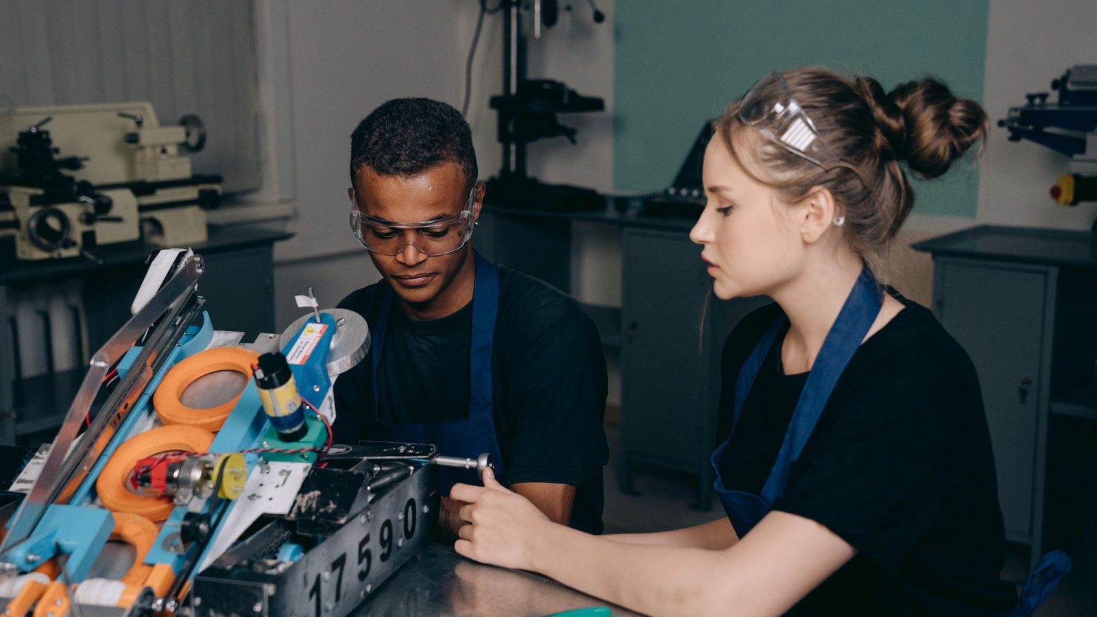 Une jeune apprentie et un jeune apprenti en tablier bleu travaillent sur un robot dans un atelier de formation