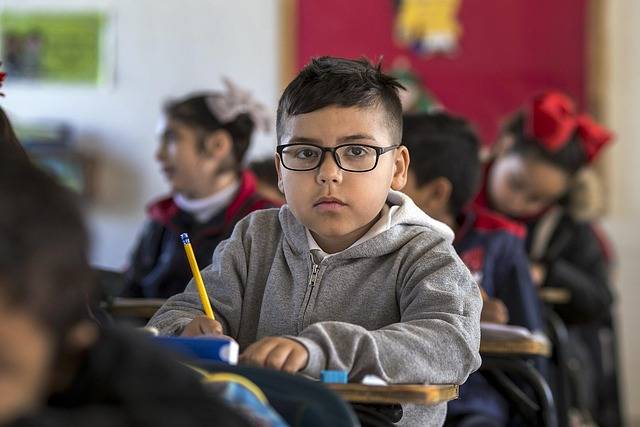 Un élève à lunettes assis à sa table en classe, crayon à la main, entouré de ses camarades