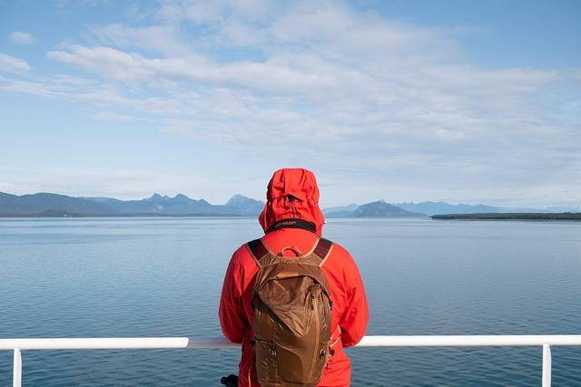 Une personne en veste rouge à capuche et sac à dos regarde la mer et les montagnes depuis le pont d'un bateau